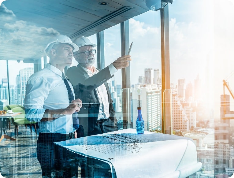 Two men in construction helmets study architectural blueprints in a sunlit office with a cityscape view. One points at a tablet, indicating planning.