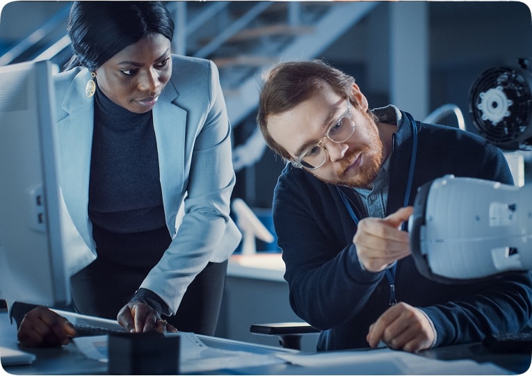 Two engineers in a modern workspace, one pointing at a device, the other observing attentively. The scene conveys focus and collaboration.