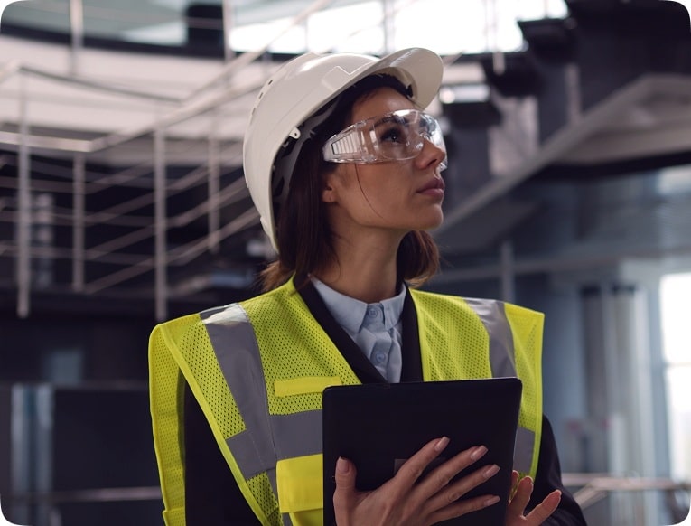 Woman in a construction site wearing a hard hat, safety glasses, and yellow vest. She holds a black tablet, looking upwards with determination.